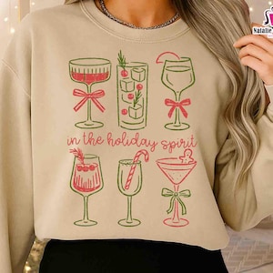 May include: Beige sweatshirt with a festive cocktail design. The graphic features six different cocktail glasses in red and green, with the phrase "in the holiday spirit" in a script font. A perfect holiday themed gift.