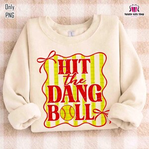 Może przedstawiać: Kremowa bluza z napisem "HIT the DANG BALL" w wytartych, czerwonych literach. Tekst umieszczono na ż&oacute;łto-białym tle w paski, z grafiką baseballu. Projekt obramowany jest czerwoną obw&oacute;dką z kokardkami.