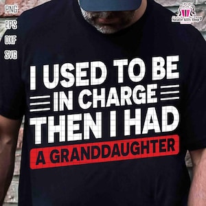 May include: Black t-shirt with white text that reads "I USED TO BE IN CHARGE THEN I HAD A GRANDDAUGHTER." The text is in a bold, sans-serif font. The words "A GRANDDAUGHTER" are in a red rectangle.