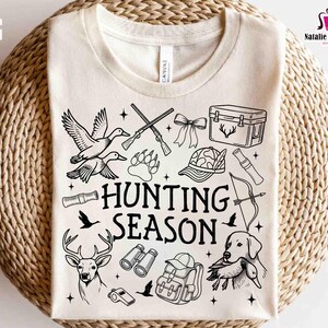 May include: Cream-colored t-shirt with a black and white hunting-themed graphic. The design includes ducks, rifles, a deer, a dog, and the words "HUNTING SEASON". The shirt also has the text "PNG EPS DXF SVG".