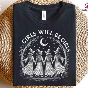 May include: Black t-shirt with the text "GIRLS WILL BE GIRLS" in white. The design features four witch figures holding hands under a crescent moon and stars. The shirt is made of a soft material.
