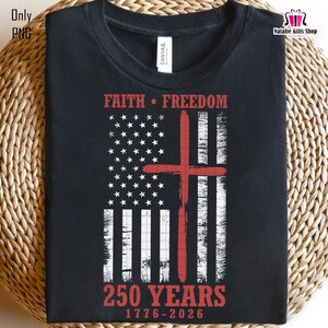 Op de afbeelding: Zwart T-shirt met een Amerikaans vlagontwerp en een rood kruis. De tekst luidt "FAITH FREEDOM" boven de vlag en "250 YEARS 1776-2026" eronder. Het T-shirt heeft een ronde hals.