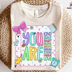Puede incluir: Una camiseta color crema con un gr&aacute;fico colorido que dice "YOU ARE" en letras grandes y divertidas. El dise&ntilde;o incluye un lazo rosa, l&aacute;pices, flores y las palabras "KIND, BRAVE, SMART, HELPFUL, GRATEFUL, ENOUGH, STRONG, UNIQUE, LOVE."