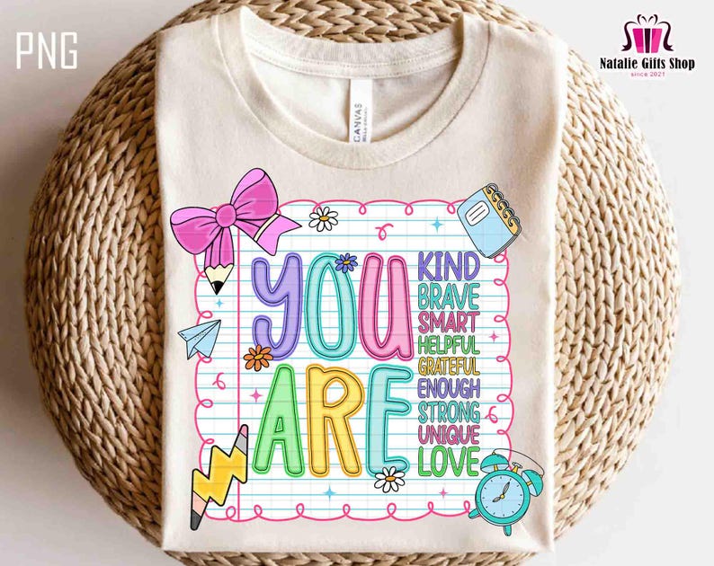Puede incluir: Una camiseta color crema con un gr&aacute;fico colorido que dice "YOU ARE" en letras grandes y divertidas. El dise&ntilde;o incluye un lazo rosa, l&aacute;pices, flores y las palabras "KIND, BRAVE, SMART, HELPFUL, GRATEFUL, ENOUGH, STRONG, UNIQUE, LOVE."