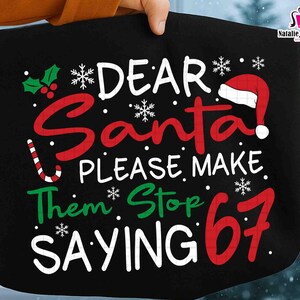 May include: Black tote bag with the text "DEAR Santa! PLEASE MAKE Them Stop SAYING 67" in white, red, and green. Includes a Santa hat, snowflakes, and holly. The bag is held by a person.