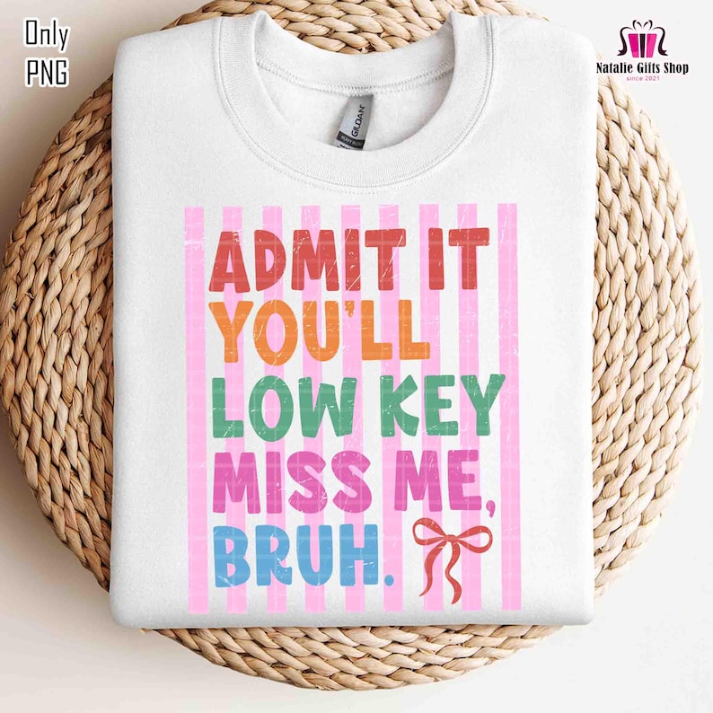 May include: White sweatshirt with the text "ADMIT IT YOU'LL LOW KEY MISS ME, BRUH." in colorful letters. The text is set against a pink striped background. A red bow is at the bottom right. The sweatshirt is on a woven surface.
