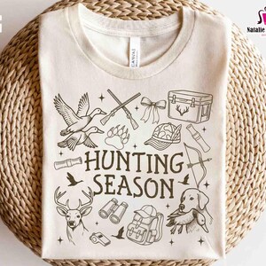May include: Cream-colored t-shirt featuring a black and white hunting-themed graphic. The design includes illustrations of ducks, rifles, a deer, a dog, and the words "HUNTING SEASON". The shirt is displayed on a woven surface.