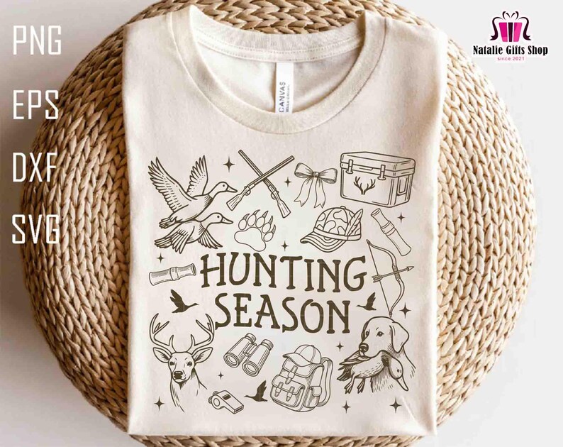 May include: Cream-colored t-shirt featuring a black and white hunting-themed graphic. The design includes illustrations of ducks, rifles, a deer, a dog, and the words "HUNTING SEASON". The shirt is displayed on a woven surface.