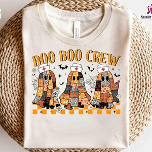 K&ouml;nnte beinhalten: Cremefarbenes T-Shirt mit dem Text "BOO BOO CREW" in Orange. Das Design zeigt drei Geisterfiguren in Patchworkmustern, die Krankenschwesternh&uuml;te tragen und medizinische Gegenst&auml;nde halten. Flederm&auml;use, Spinnweben und Blumen vervollst&auml;ndigen das Halloween-Thema.