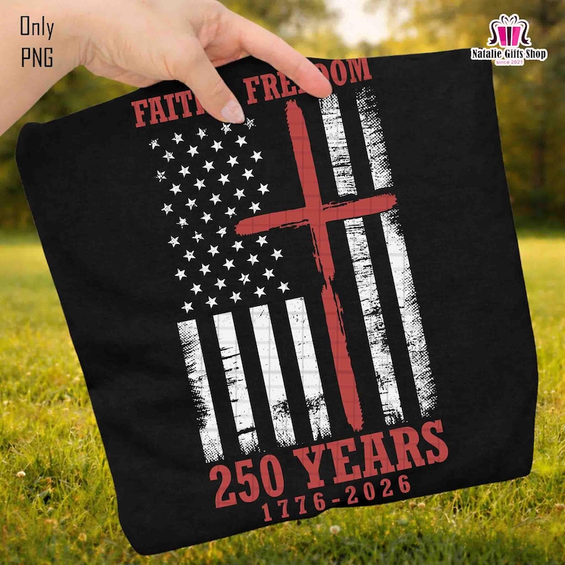 Op de afbeelding: Zwarte stof met een verweerd Amerikaans vlagontwerp, met een rood kruis eroverheen. De woorden "FAITH FREEDOM" en "250 YEARS 1776-2026" zijn in het rood gedrukt. De stof wordt tegen een achtergrond van groen gras gehouden.