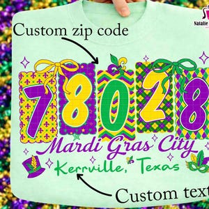 Op de afbeelding: Een lichtgroene sweatshirt met een Mardi Gras-thema. Het ontwerp bevat de nummers 78028 in geel en paars, met de tekst "Mardi Gras City Kerrville, Texas". Het ontwerp bevat ook een hoed, masker en strik.