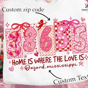 Może przedstawiać: Biała bluza z motywem walentynkowym. Zawiera niestandardowy projekt kodu pocztowego w kolorze różowym i czerwonym, z napisem "HOME IS WHERE THE LOVE IS" i "Oxford.mississippi". Zawiera kokardy i serca.