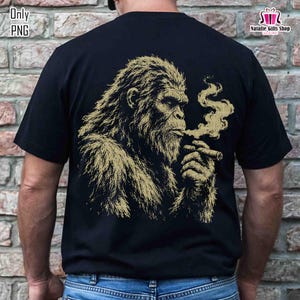 Può includere: T-shirt nero con un'illustrazione dorata di un Bigfoot che fuma un sigaro. Il disegno mostra il volto e la parte superiore del corpo del Bigfoot, con fumo che si alza dal sigaro. La maglietta ha un girocollo classico e maniche corte.