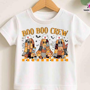 K&ouml;nnte beinhalten: Wei&szlig;es T-Shirt mit dem orangefarbenen Text "BOO BOO CREW". Das Design zeigt drei Geisterfiguren in Patchwork-Mustern, die Krankenschwesternh&uuml;te tragen und medizinische Gegenst&auml;nde halten. Flederm&auml;use und Spinnweben vervollst&auml;ndigen das Halloween-Thema.