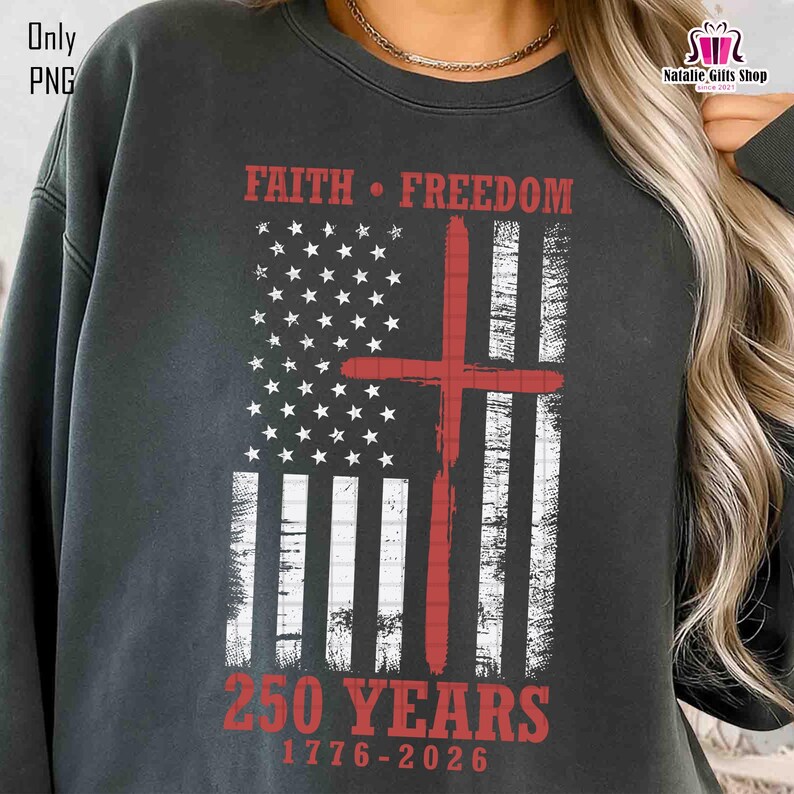 Op de afbeelding: Donkergrijze sweatshirt met een verweerd Amerikaans vlagontwerp en een rood kruis. De tekst luidt "FAITH &bull; FREEDOM" en "250 YEARS 1776-2026". De sweatshirt heeft een ronde hals.