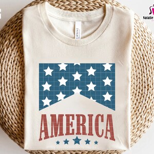 May include: A white t-shirt with a blue and white American flag design and the word "AMERICA" in red letters. There are five small stars below the word "AMERICA".