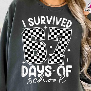 May include: Dark grey sweatshirt with the text "I Survived 67 Days of School" in white. The numbers "67" are filled with a black and white chequered pattern. The sweatshirt also has the text "DAYS OF school" in a script font.