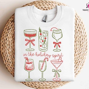 May include: White sweatshirt featuring a festive design of cocktail glasses with red and green accents. The phrase "in the holiday spirit" is printed below the glasses. The sweatshirt is displayed on a woven, circular placemat.