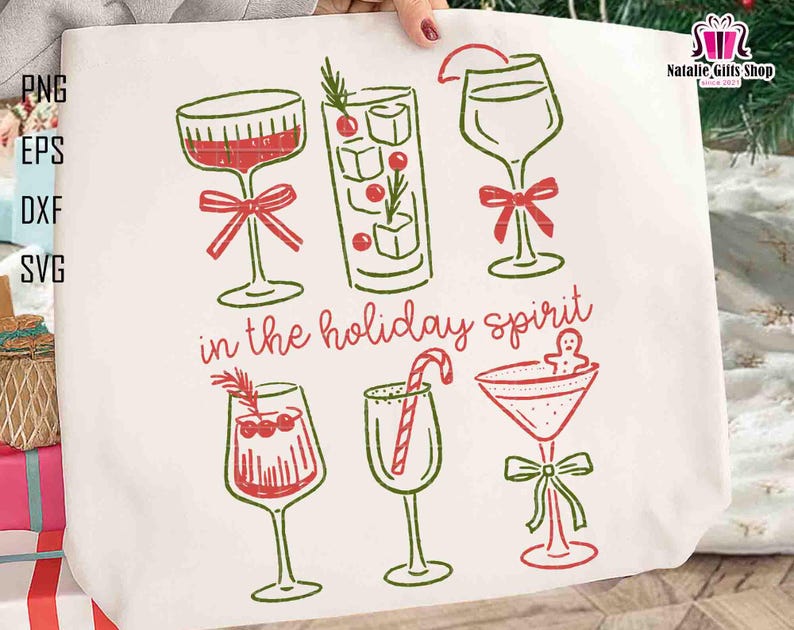 May include: Off-white tote bag featuring a festive design with six different cocktail glasses in red and green. The phrase "in the holiday spirit" is written in cursive. The bag has a simple, line-art style.