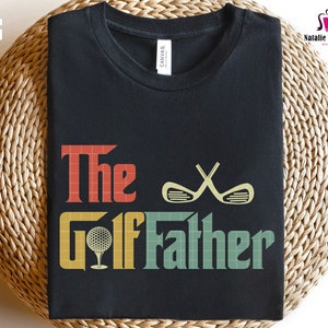 Op de afbeelding: Een zwart T-shirt met de tekst "The Golf Father" in een retro stijl met een golfbal en gekruiste golfclubs.