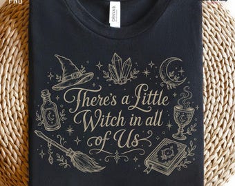 May include: A black t-shirt featuring a gold-toned graphic print with the text "There's a Little Witch in all of Us." The design includes illustrations of a witch hat, crystals, a crescent moon, a potion bottle, a broom, and a spellbook.