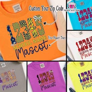 May include: A collection of crew neck t-shirts in various colors, each featuring a custom school design. The design includes the numbers "12345", the school name, and the word "Mascot". The shirts are made of a soft material.