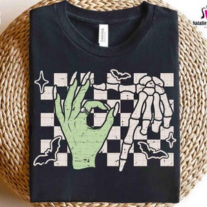 May include: Black t-shirt featuring a green hand with a ring making an 'okay' gesture, paired with a skeleton hand. The design includes bats, stars, and a checkered background. The shirt also has the text "CANVAS".