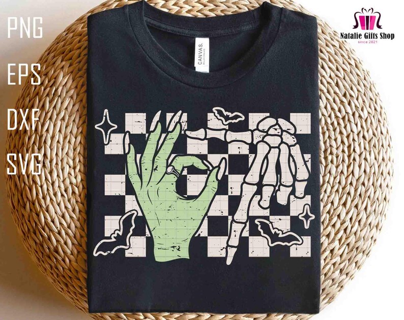 May include: Black t-shirt featuring a green hand with a ring making an 'okay' gesture, paired with a skeleton hand. The design includes bats, stars, and a checkered background. The shirt also has the text "CANVAS".