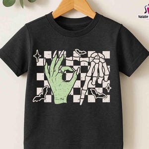 May include: Black t-shirt featuring a green hand making an 'okay' gesture and a skeleton hand, set against a checkered background with bats and stars. The shirt has the text 'PNG, EPS, DXF, SVG' on the left side.