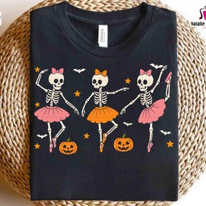 Puede incluir: Camiseta negra con un dise&ntilde;o de Halloween de tres bailarinas esqueleto con tut&uacute;s rosas y naranjas, lazos y zapatillas de ballet. El dise&ntilde;o incluye murci&eacute;lagos, estrellas y calabazas. La camiseta tiene una etiqueta "CANVAS".