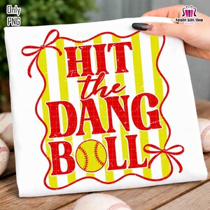May include: White t-shirt with a red and yellow design. The design features the text "HIT the DANG BALL" in red, with a yellow softball graphic. Yellow and white vertical stripes are in the background. Red bows are at the top and bottom.