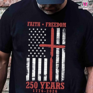 Op de afbeelding: Zwart t-shirt met een verweerd Amerikaans vlagontwerp en een rood kruis. De woorden "FAITH &bull; FREEDOM" staan boven de vlag en "250 YEARS 1776-2026" staan eronder. Het ontwerp is gedrukt op een t-shirt.