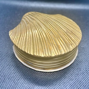 May include: A gold-colored, seashell-shaped trinket box. The lid is textured with radiating lines, mimicking the natural ridges of a seashell. The base of the box has a series of horizontal lines. The box is made of metal.