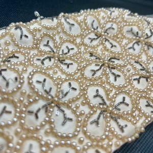 May include: Close-up of a cream-colored fabric with a delicate pattern of small, white pearls and silver beads. The beads are arranged in a repeating floral design.