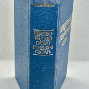 May include: A blue hardcover book with gold lettering. The spine displays the title "NEMECKO-RUSSKIJ RUSSKO-NEMECKII SLOVARB" in gold. The pages are slightly yellowed, indicating age. The book appears to be a German-Russian dictionary.