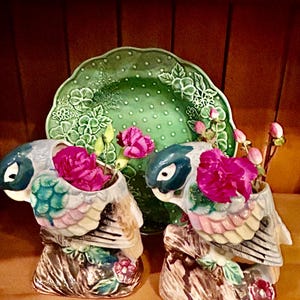May include: Two ceramic parrot-shaped vases with pink flowers, set in front of a green, floral-patterned plate. The vases are perched on faux tree stumps, with blue, white, and pastel-colored wings. The plate has a raised floral design.