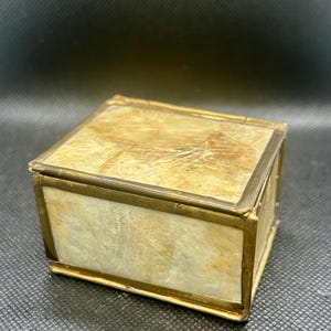 May include: A small, square box with a light-colored, marbled top and sides. The box is trimmed with a thin, gold-colored metal. The box is likely used for storing small items.