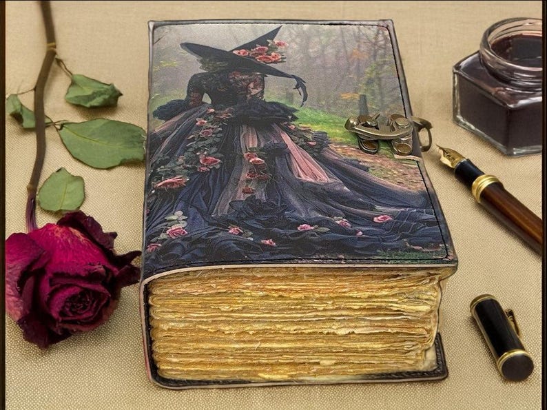 Puede incluir: Un diario decorativo con una ilustraci&oacute;n de bruja en la portada, con un vestido negro con rosas rosas. El diario tiene p&aacute;ginas envejecidas y amarillentas y un cierre de metal. Una rosa seca y un tintero est&aacute;n cerca.