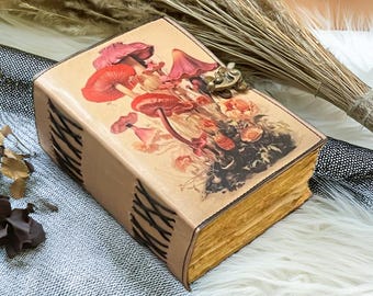 May include: A tan leather-bound journal with a colorful mushroom illustration on the cover. The journal has a decorative clasp and stitched binding. The pages have a vintage, aged appearance. A unique, handcrafted item.