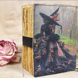 Puede incluir: Un diario decorativo con una portada tem&aacute;tica de brujas. La portada presenta a una mujer con un vestido negro y un sombrero de bruja adornado con rosas. El diario tiene p&aacute;ginas envejecidas y una banda negra. Una rosa seca est&aacute; al lado.
