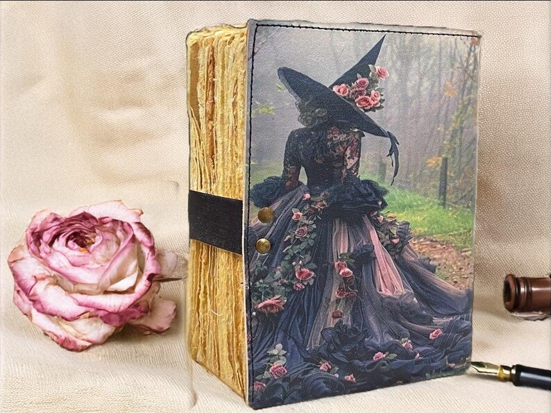 Puede incluir: Un diario decorativo con una portada tem&aacute;tica de brujas. La portada presenta a una mujer con un vestido negro y un sombrero de bruja adornado con rosas. El diario tiene p&aacute;ginas envejecidas y una banda negra. Una rosa seca est&aacute; al lado.