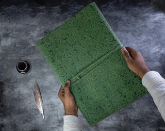 May include: A green journal with a detailed floral design is held open. A black inkwell and a feather quill are next to the journal on a dark, textured surface. The journal's cover is a deep green colour.