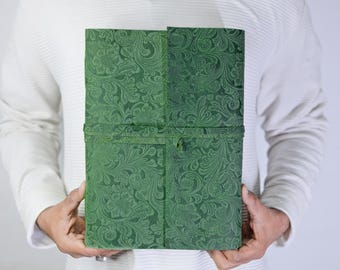 May include: A green journal with a floral embossed pattern. The journal is held open, revealing the inside pages. The cover is a deep green colour, and the floral design is visible across the entire surface. The journal is secured with a matching green strap.
