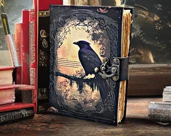 Può includere: Un diario rilegato in pelle scura con un'illustrazione di un corvo. La copertina presenta una cornice ornata attorno al corvo appollaiato su un ramo. Il diario è chiuso con un cinturino in pelle nera e una chiusura in metallo. Diversi libri rossi sono impilati dietro il diario.