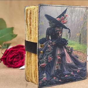Puede incluir: Un diario hecho a mano con una cubierta decorativa que presenta a una mujer con un disfraz de bruja con un sombrero grande y adornos florales. El diario tiene una correa negra y remaches de lat&oacute;n. Una rosa roja y vegetaci&oacute;n est&aacute;n en el fondo.