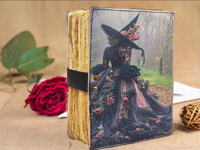 Puede incluir: Un diario hecho a mano con una cubierta decorativa que presenta a una mujer con un disfraz de bruja con un sombrero grande y adornos florales. El diario tiene una correa negra y remaches de lat&oacute;n. Una rosa roja y vegetaci&oacute;n est&aacute;n en el fondo.