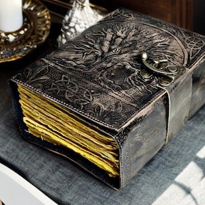 May include: An antique-style journal with a tree of life design embossed on the cover. The journal has a metal clasp, a leather strap, and aged, yellowed pages. The cover is dark grey with intricate Celtic knotwork.