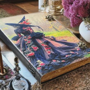 Puede incluir: Un diario decorativo con una portada de arte fant&aacute;stico que presenta a una bruja con un vestido fluido adornado con rosas. El diario tiene una lomo negro, cierres met&aacute;licos y p&aacute;ginas envejecidas. La portada muestra una bruja en un bosque.