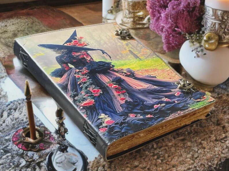 Puede incluir: Un diario decorativo con una portada de arte fant&aacute;stico que presenta a una bruja con un vestido fluido adornado con rosas. El diario tiene una lomo negro, cierres met&aacute;licos y p&aacute;ginas envejecidas. La portada muestra una bruja en un bosque.