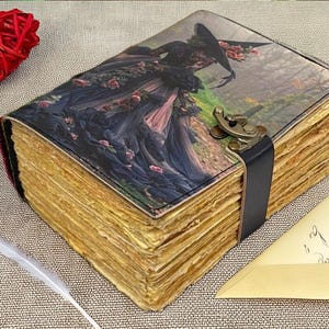 Puede incluir: Un diario de estilo vintage con una ilustraci&oacute;n g&oacute;tica en la portada, que representa a una mujer con un vestido negro y un sombrero. El diario tiene p&aacute;ginas amarillentas envejecidas, una correa de cuero negro y un cierre de metal. Un coraz&oacute;n rojo y rosas secas son visibles.